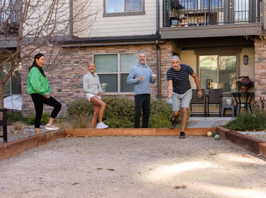 Four people playing bocce on a gravel court outside brick apartment buildings