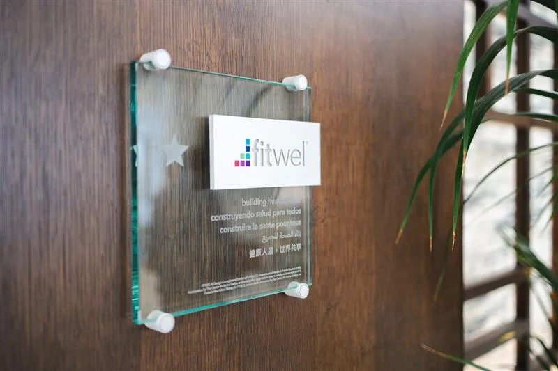 A glass Fitwel plaque with text in multiple languages is mounted on a wooden wall at Columbus Library, partially framed by green plant leaves on the right side.