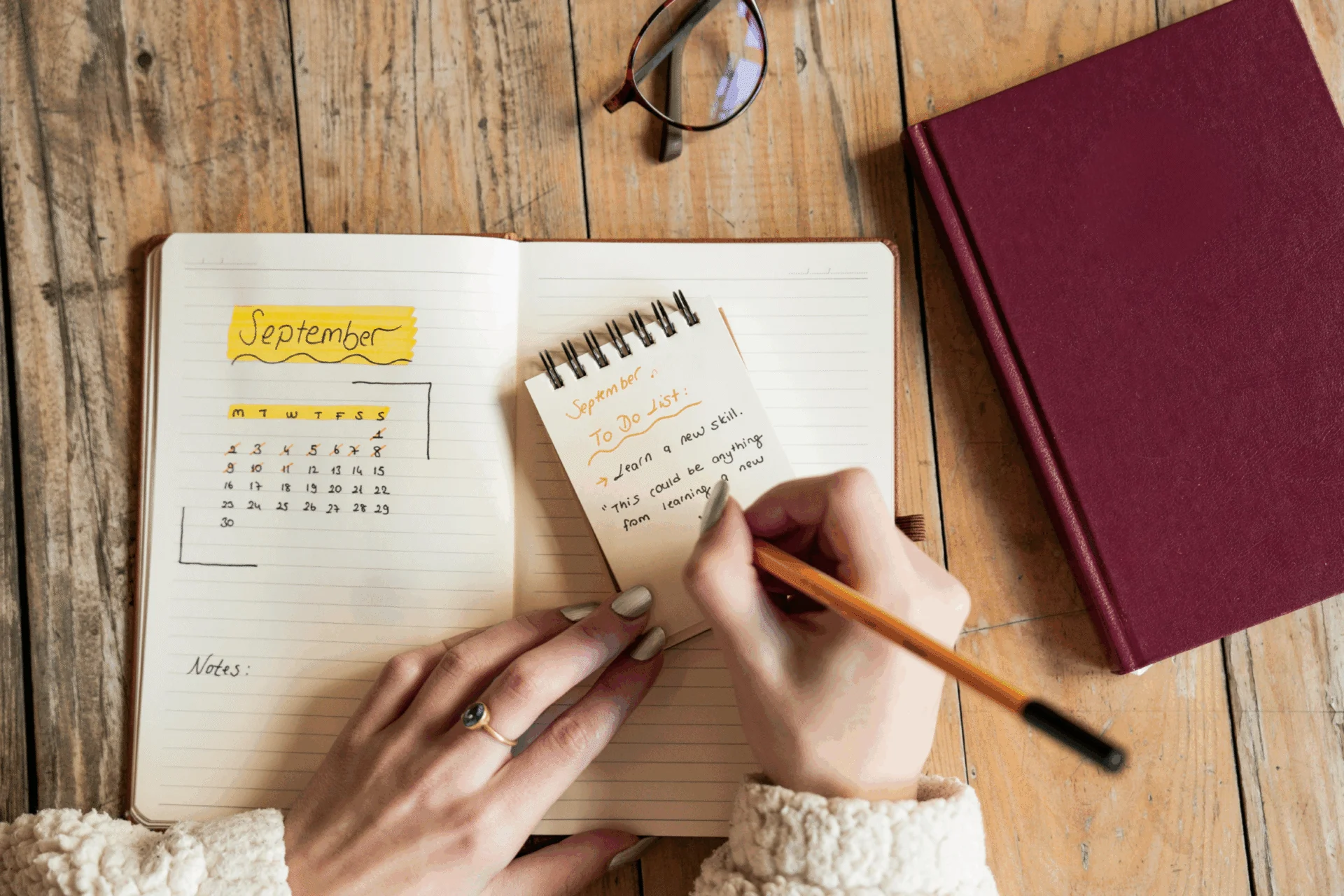 Hands writing in a notebook beside a monthly calendar on a wooden desk