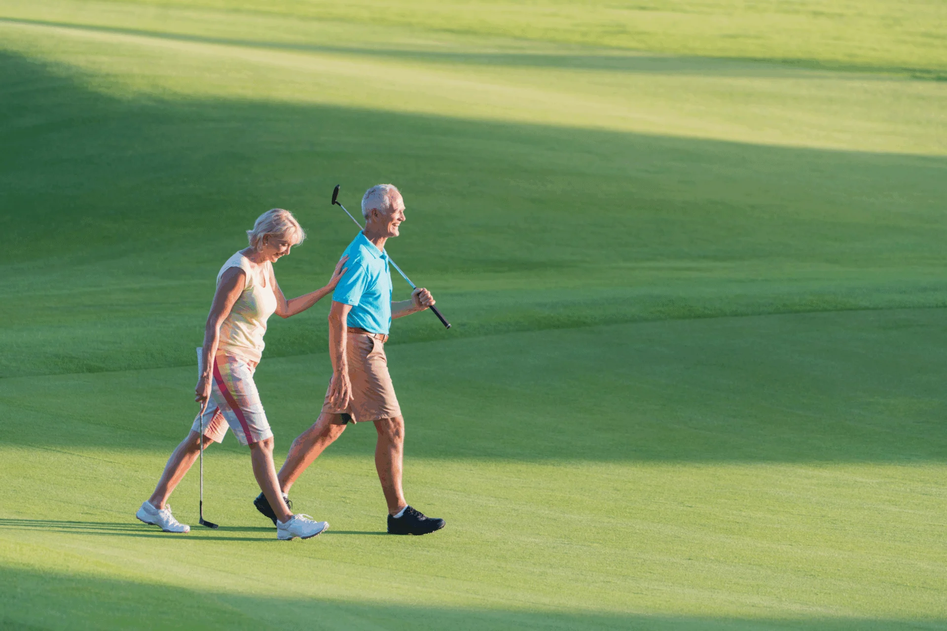 A senior couple strolls side by side on a sunny, luxury golf course in Hilliard Ohio, each holding golf clubs with green grass and long shadows stretching behind them.